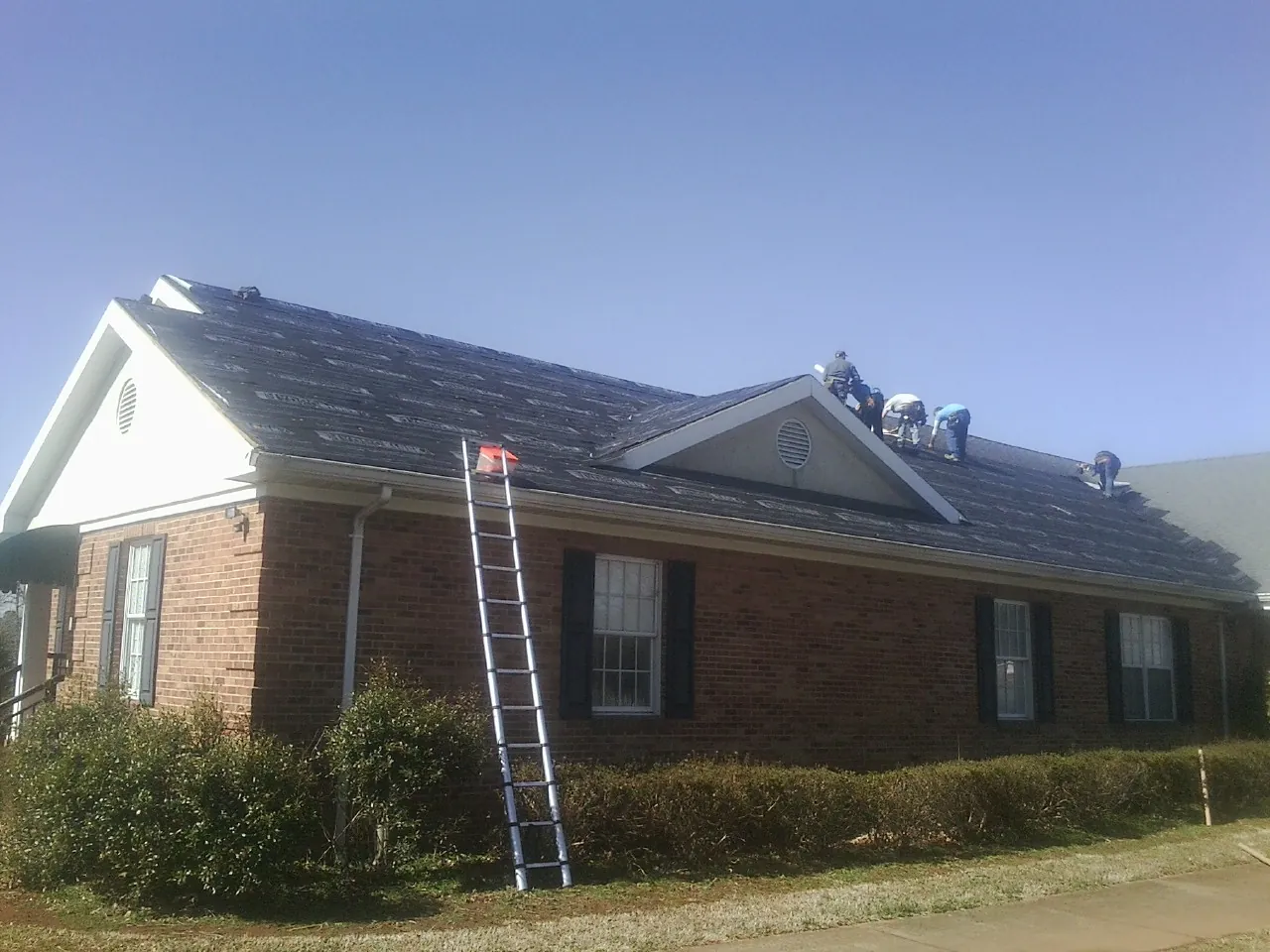 Crew tearing shingles off a brick ranch roof with a ladder set — Metal Roof Repair work by our crew in Delevan