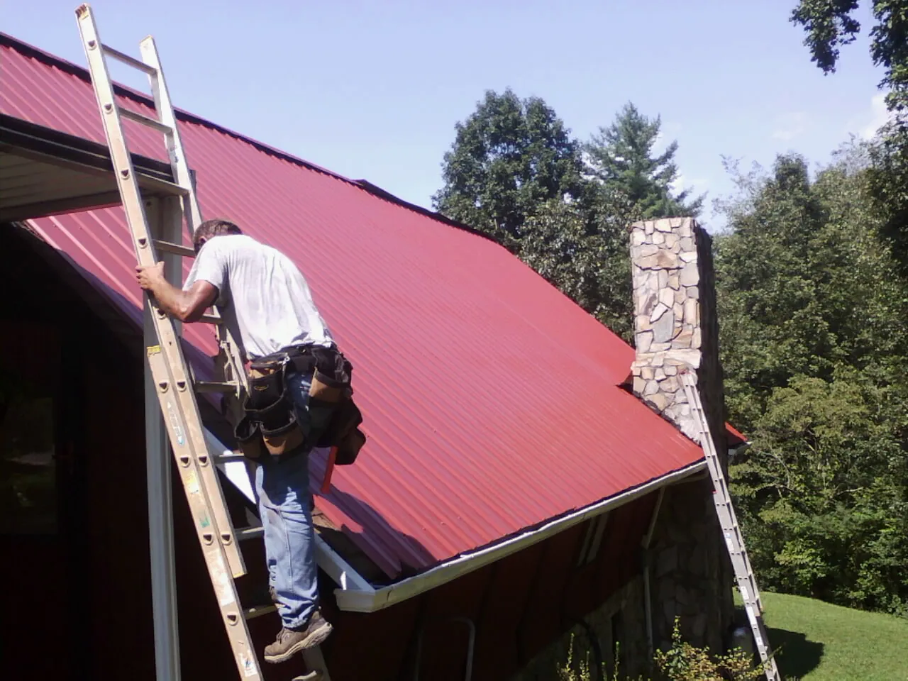 Roofer on a ladder beside a red metal roof and stone chimney — Soffit & Fascia Repair by Roofing Brothers in Delevan